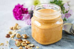 Homemade Hazelnut Butter in a glass jar next to flowers and chopped hazelnuts.