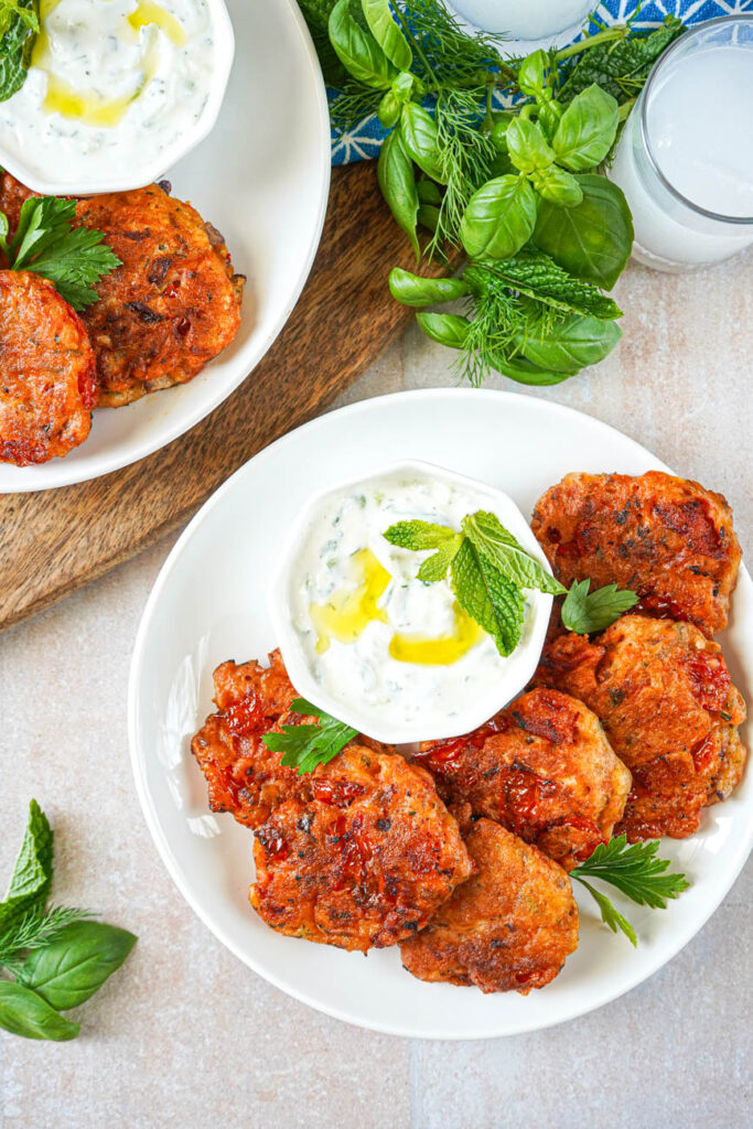 Aerial view of Santorini Tomato Patties (Tomatokeftedes Santorinis) on two white plates next to two glasses of ouzo and a bundle of herbs.