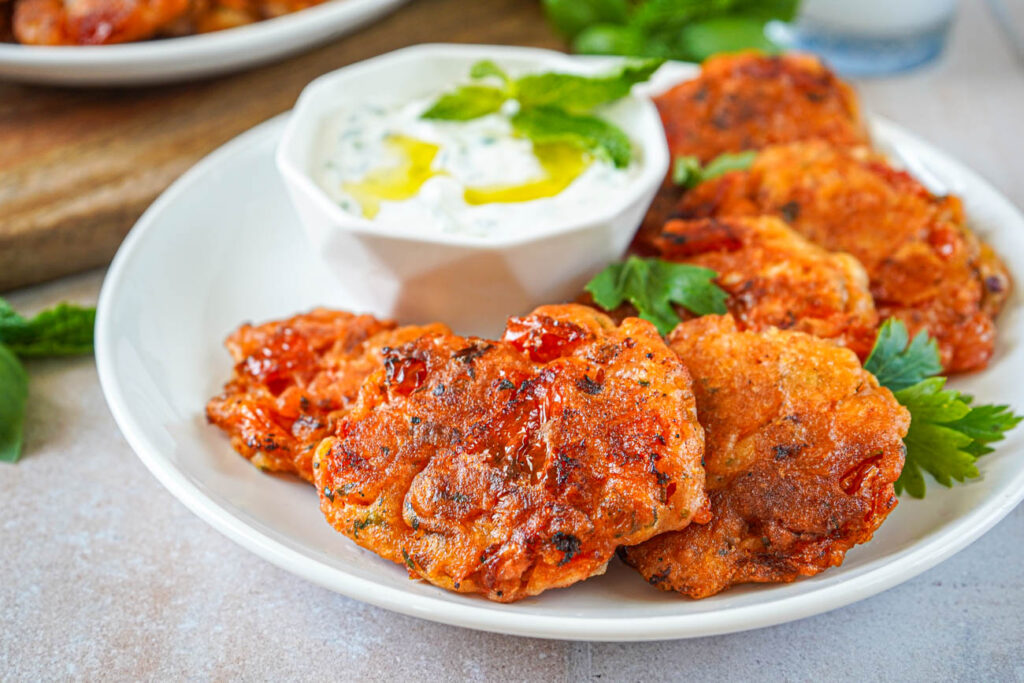 Santorini Tomato Patties (Tomatokeftedes Santorinis) on a white plate with parsley leaves next to a bowl of tzatziki.