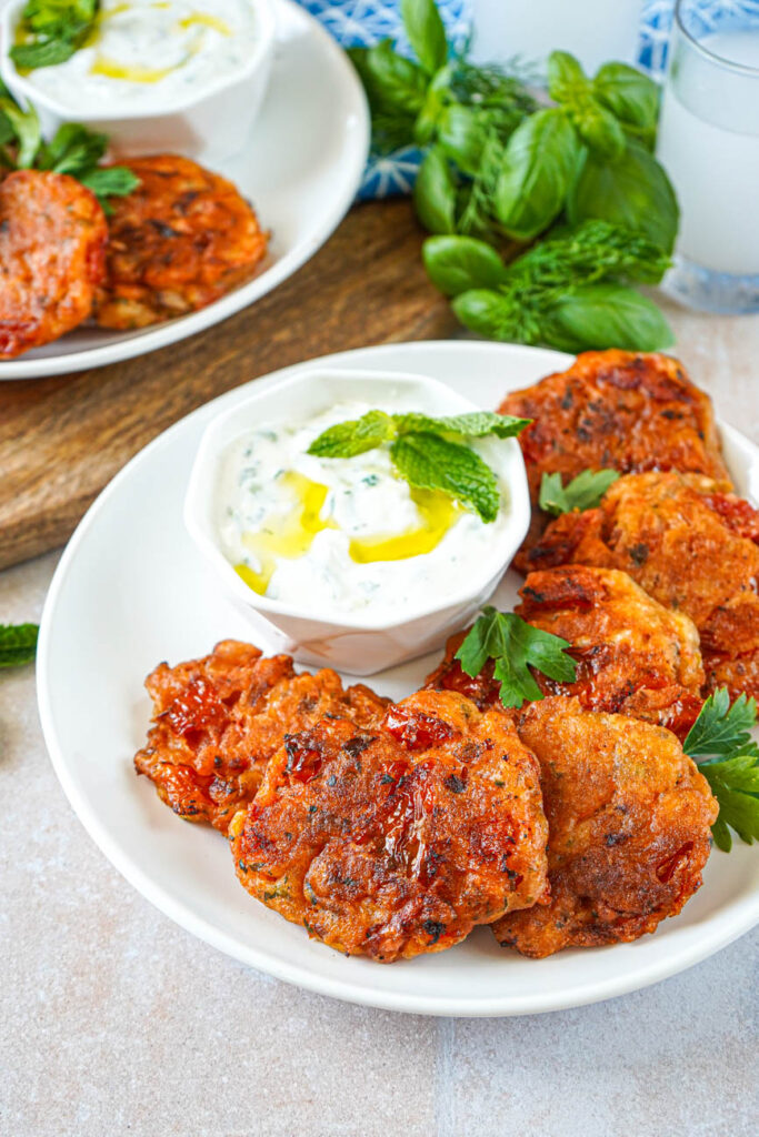 Close up of Santorini Tomato Patties (Tomatokeftedes Santorinis) on a white plate with another plate in the background, herbs, and two glasses.