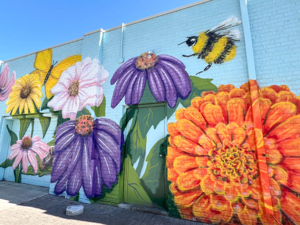 Mural of orange, purple, pink, and yellow flowers with a bumble bee and butterfly on a brick wall.