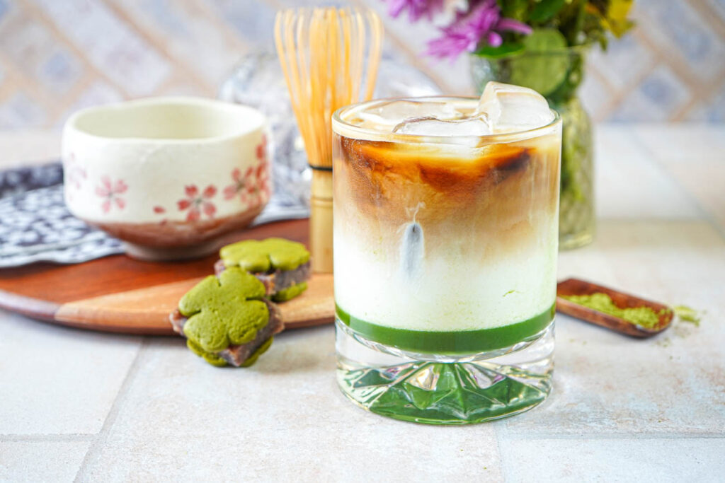 Side view of Iced Matcha Espresso Latte in a glass with a vase of flowers, whisk, and bowl in the background.