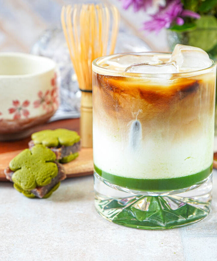 Iced Matcha Espresso Latte in a glass with ice next to two matcha yokan cookies, a bamboo whisk, and a cherry blossom bowl.