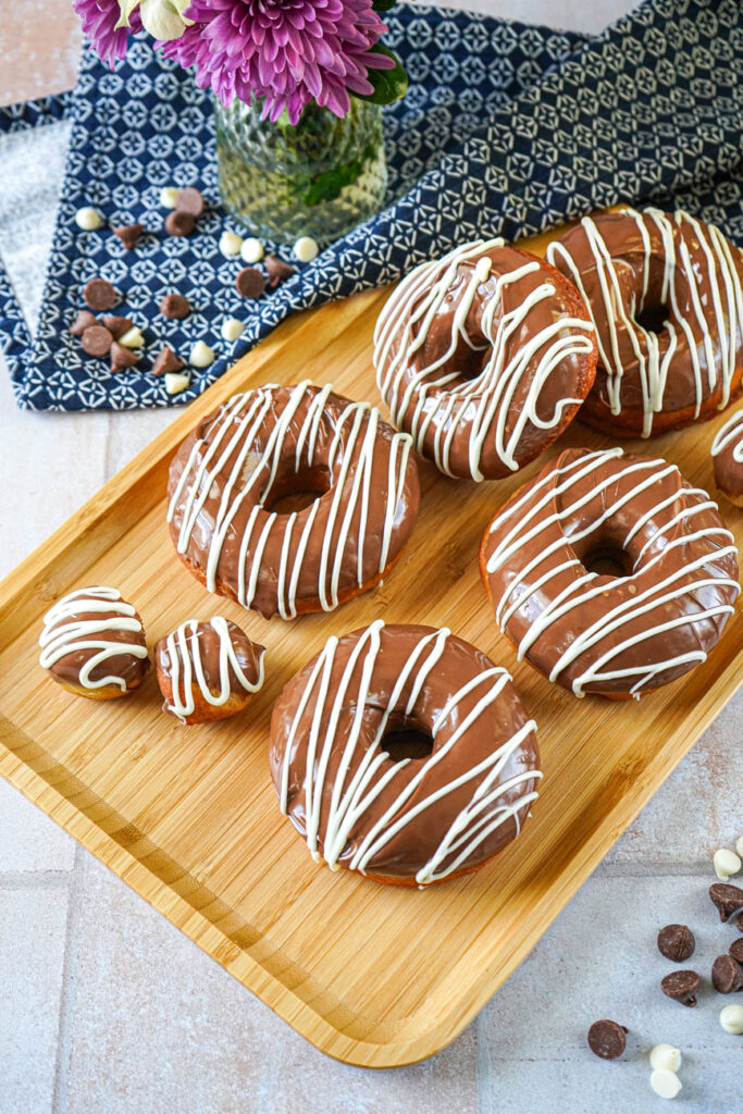 Aerial view of Milk Chocolate Donuts and donut holes on a wooden tray next to chocolate chips and a vase with flowers.