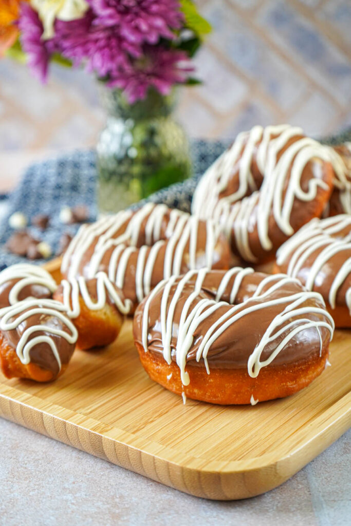 Side view of Milk Chocolate Donuts on a wooden tray with a vase of flowers in the background.