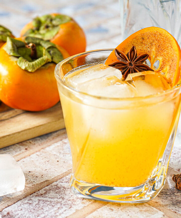 Kaki Sour in a glass with ice, a persimmon slice, and star anise.