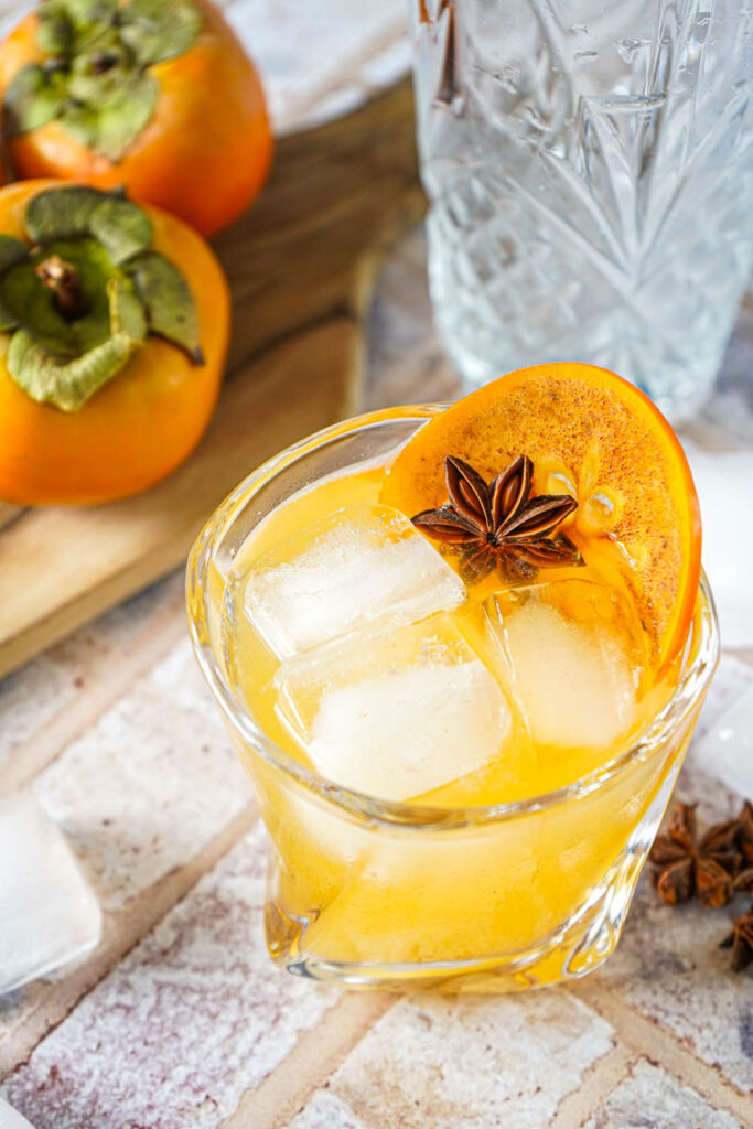 Close up of Kaki Sour in a glass with a persimmon slice, ice, and a star anise.