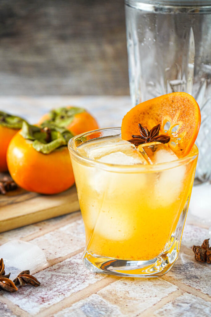 Side view of Kaki Sour in a glass with persimmons and a cocktail shaker in the background.