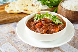 Lamb and Potato Curry in a white bowl with rice and bread in the background.