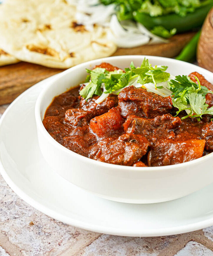 Lamb and Potato Curry in a white bowl with rice and bread in the background.