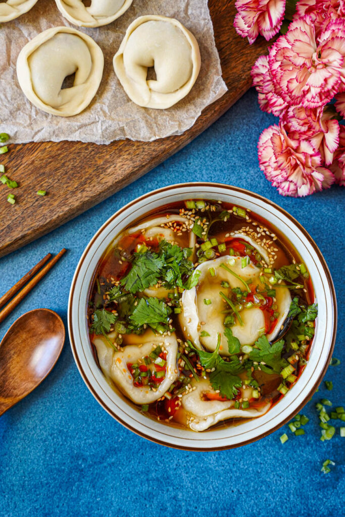 Aerial view of Shaanxi Dumpling Soup in a bowl next to pink flowers and dumplings on a sheet of parchment paper.