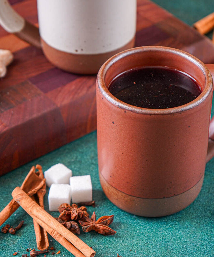 Feuerzangenbowle (German Fire Tongs Punch) in a red mug next to cinnamon sticks, star anise, and sugar cubes.