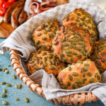 Kürbiskernbrötchen (Pumpkin Seed Rolls) in a basket with sausage, cheese, and grape tomatoes in the background.