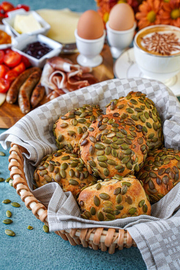 Kürbiskernbrötchen (Pumpkin Seed Rolls) in a basket with coffee, eggs, Speck, and sausage in the background.