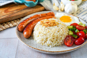 Silog- Garlic fried rice and a fried egg on a plate with two hot dogs and tomato cucumber salad.