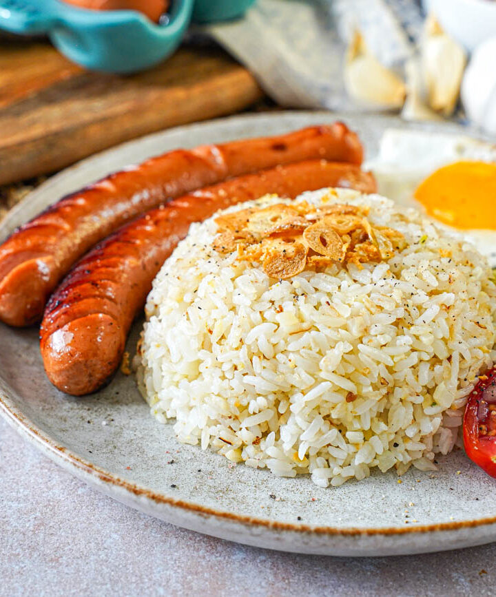 Silog- Garlic fried rice and a fried egg on a plate with two hot dogs and tomato cucumber salad.