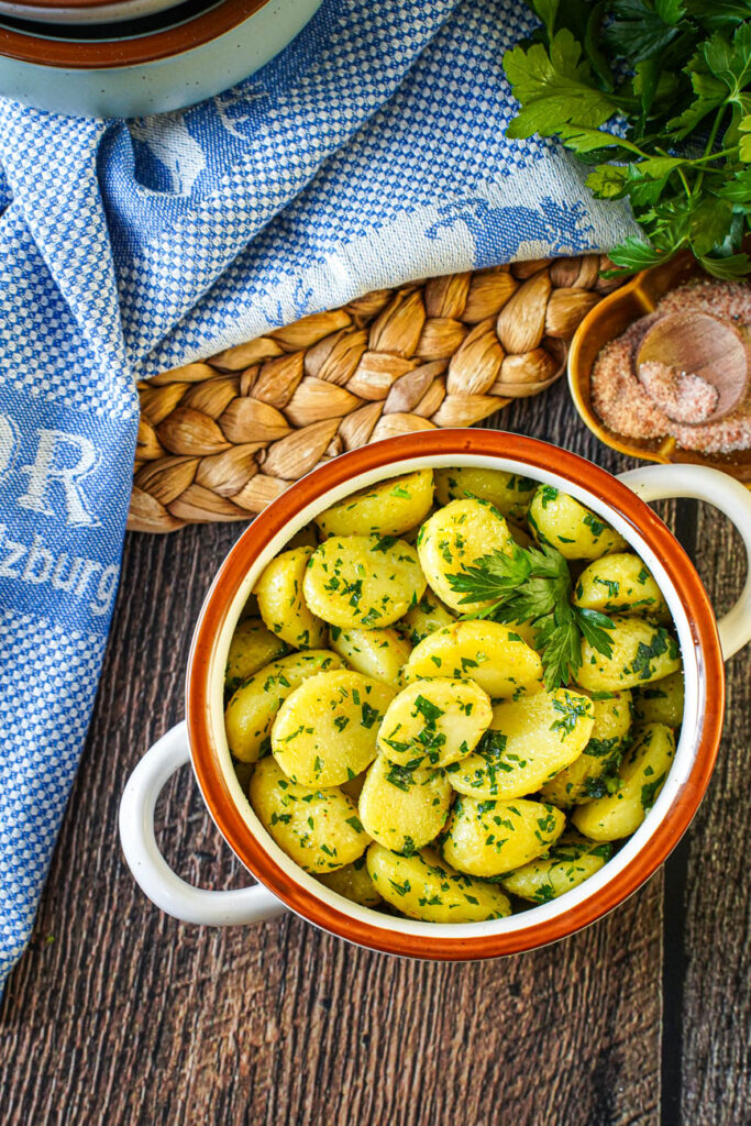 Aerial view of Petersilienerdäpfel (Parsley Potatoes) in a white bowl next to a bowl of pink salt and a blue towel.