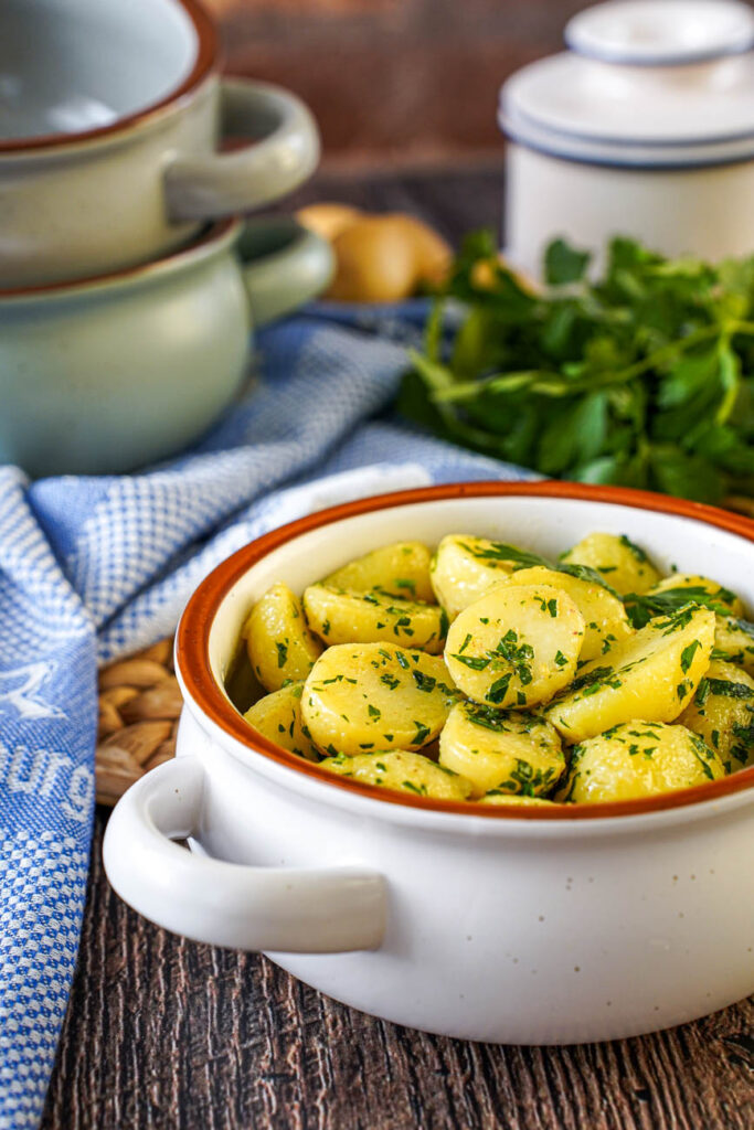 Side view of Petersilienerdäpfel (Parsley Potatoes) in a white bowl with three stacked bowls and a bunch of parsley in the background.
