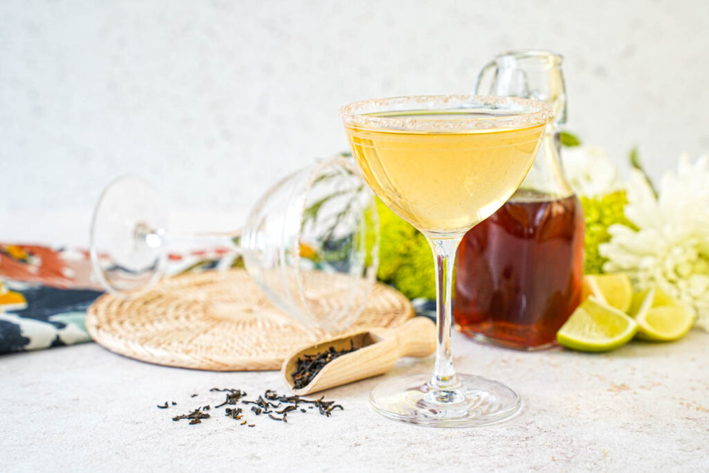 Smoky Lapsang Martini in a glass with a jar of Lapsang syrup in the background next to lime wedges and tea leaves.