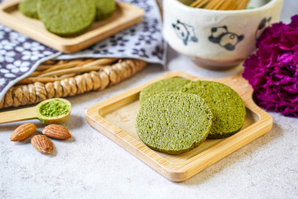 Matcha Butter Almond Cookies on a small wooden board next to almonds and a small spoon with matcha.
