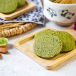 Three Matcha Butter Almond Cookies on a small wooden board next to almonds and a spoonful of matcha.