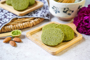 Three Matcha Butter Almond Cookies on a small wooden board next to almonds and a spoonful of matcha.