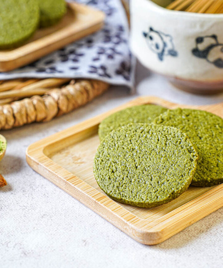 Three Matcha Butter Almond Cookies on a small wooden board next to almonds and a spoonful of matcha.