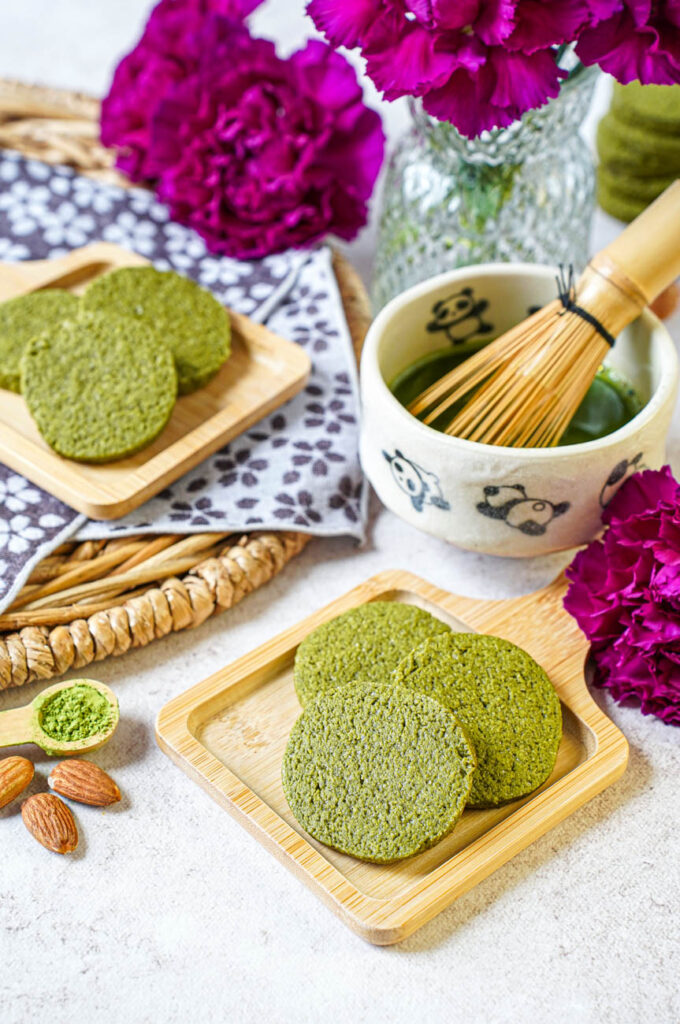 Close up of Matcha Butter Almond Cookies on two small wooden boards next to a panda matcha bowl, whisk, almonds, and purple flowers.