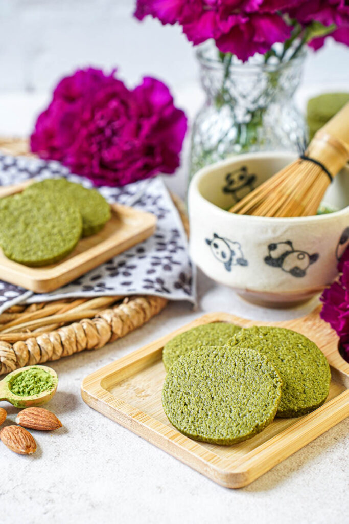 Side view of Matcha Butter Almond Cookies on two small wooden boards next to a panda matcha bowl, almonds, and purple flowers.