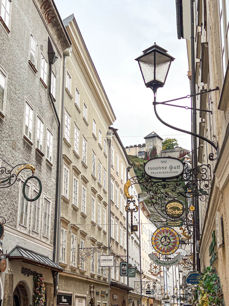 Street in Salzburg with signs outside of the buildings and a stone fortress in the background.