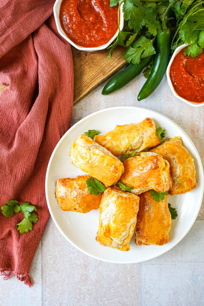 Aerial view of Paneer Masala Puffs on a plate next to cilantro leaves and a bowl of tomato chile sauce.