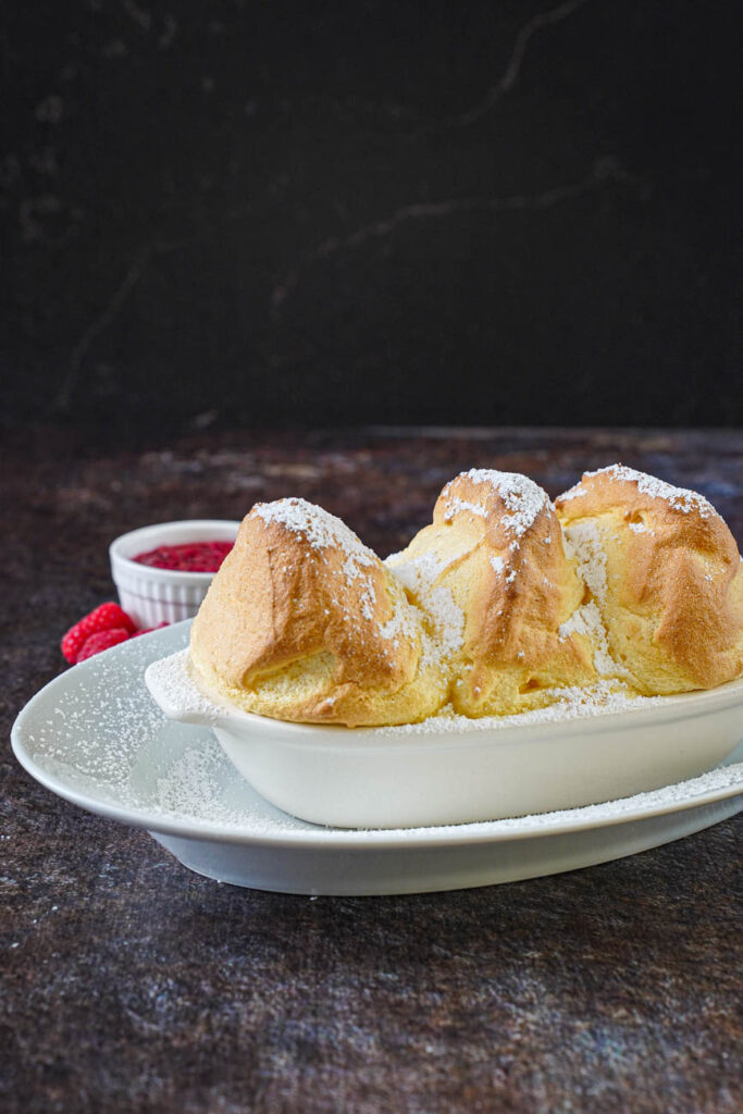 Miniature Salzburger Nockerl in a small white gratin dish with raspberry sauce and fresh raspberries in the background.