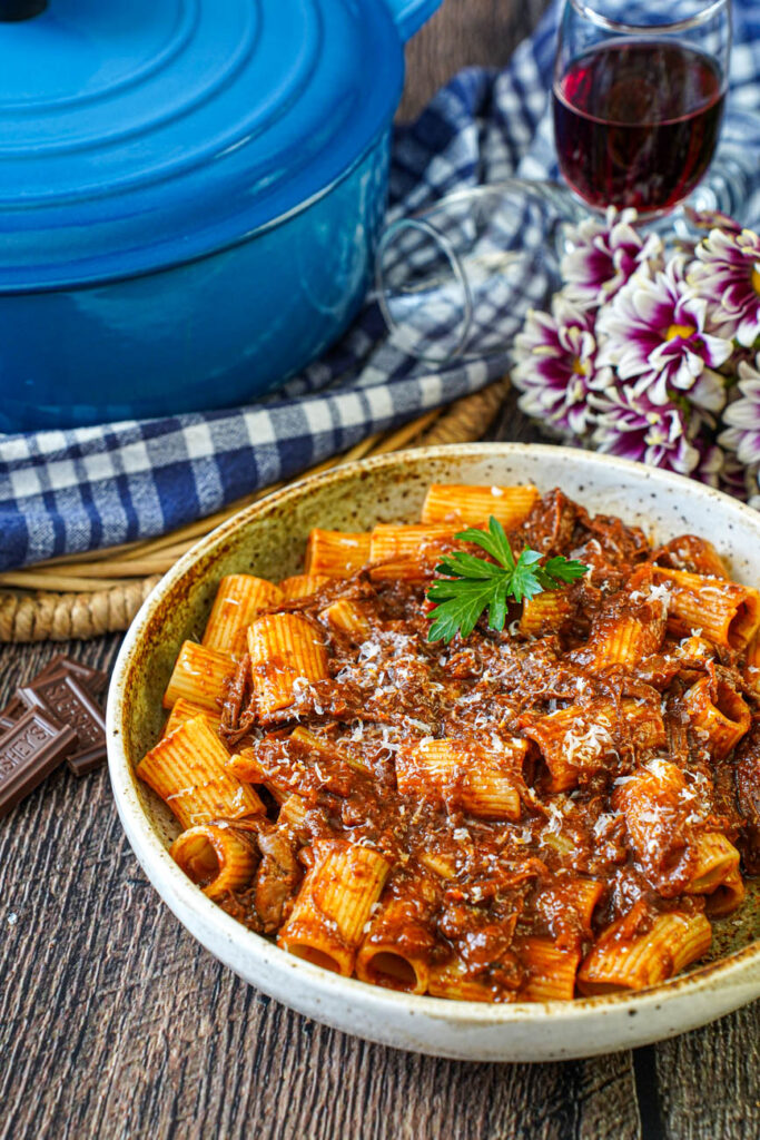 Short Rib Ragu in a bowl with rigatoni and next to purple and pink flowers, a blue Dutch oven, and three small Hershey bar pieces.