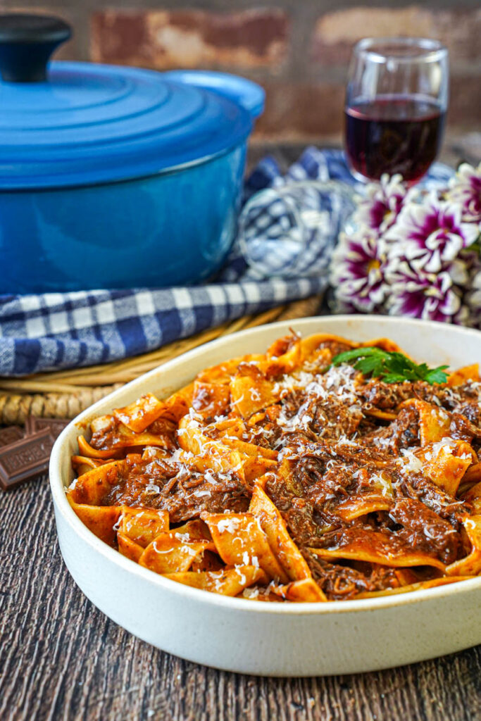 Side view of Short Rib Ragu in a bowl with pappardelle and a glass of red wine in the background next to a blue Dutch oven.