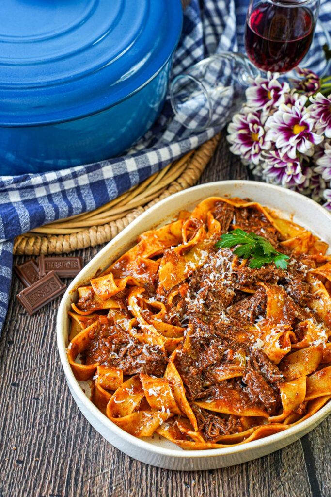Short Rib Ragu in a bowl with pappardelle and next to purple and white flowers, a glass of red wine, a blue Dutch oven, and three small chocolate pieces.