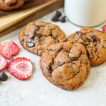 Three Strawberry Chocolate Chip Cookies next to dried strawberry pieces and chocolate chips.