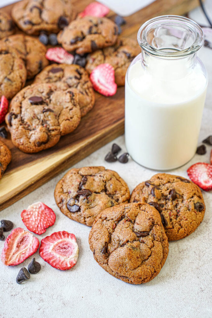Strawberry Chocolate Chip Cookies next to freeze dried strawberry slices, chocolate chips, and a glass of milk.