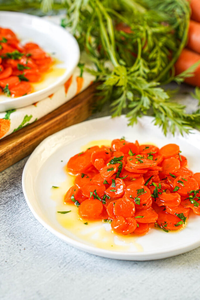 Close up view of Vichy Carrots (Carottes Vichy) on two plates with a bunch of carrots and carrot leaves in the background.