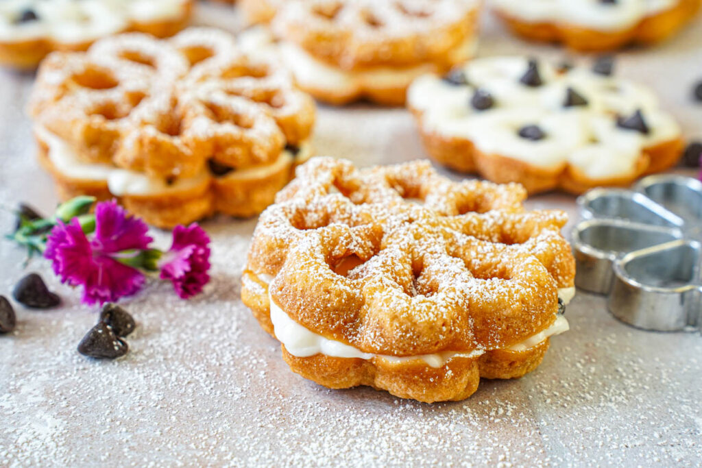 Baci Panteschi (Flower-Shaped Ricotta-Filled Pastries) filled with a sweet ricotta cream next to pink flowers and chocolate chips.