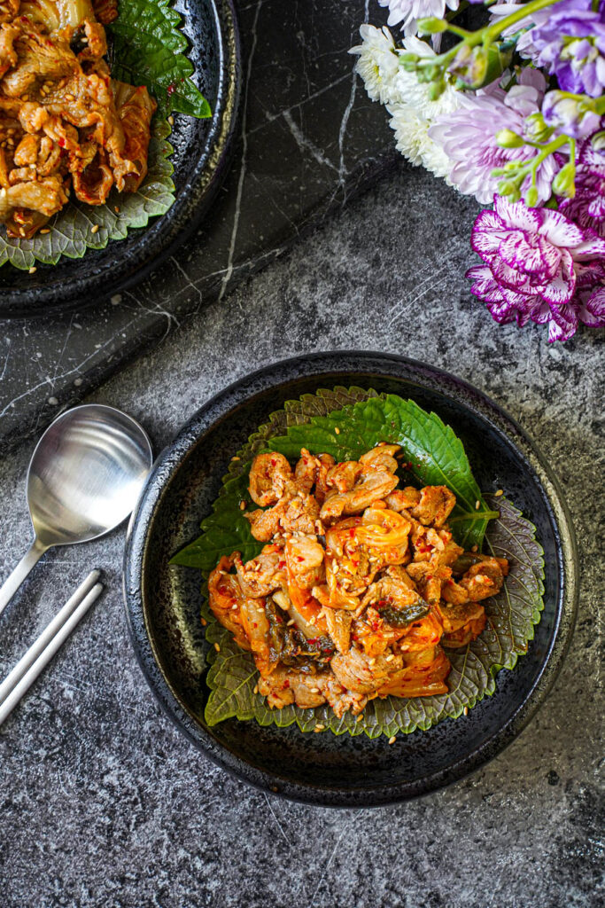 Aerial view of Pork and Kimchi Stir-Fry in two black bowls next to purple flowers.