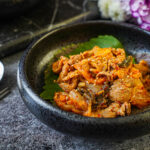 Pork and Kimchi Stir-Fry in a black bowl next to a metal spoon and chopsticks.