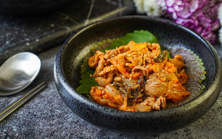 Pork and Kimchi Stir-Fry in a black bowl next to a metal spoon and chopsticks.