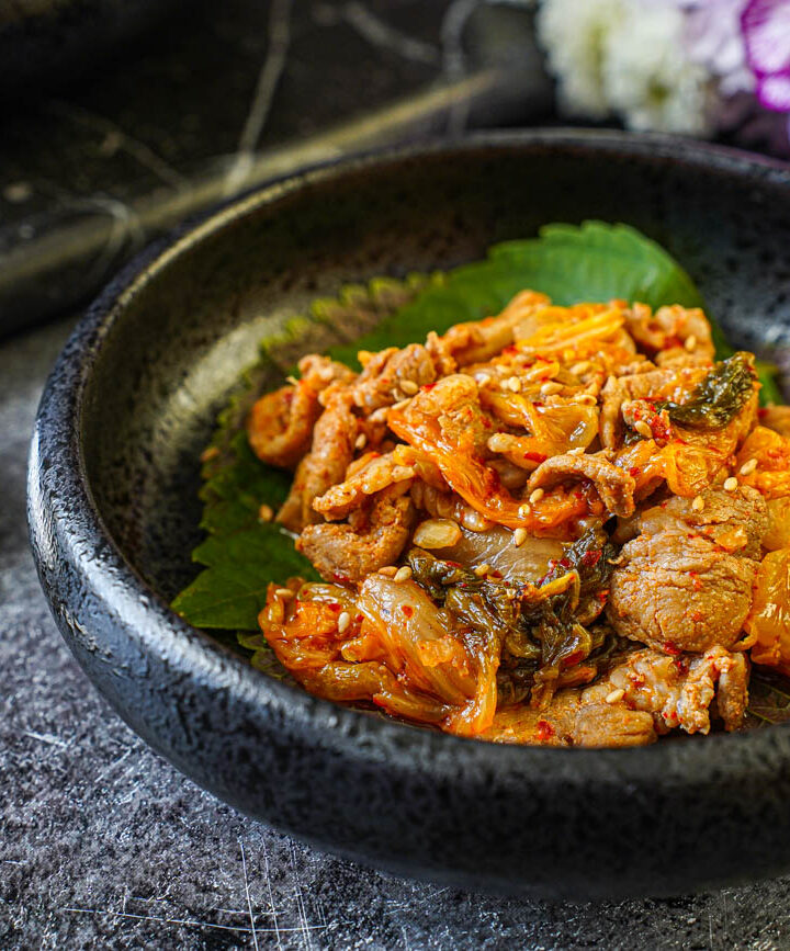 Pork and Kimchi Stir-Fry in a black bowl next to a metal spoon and chopsticks.