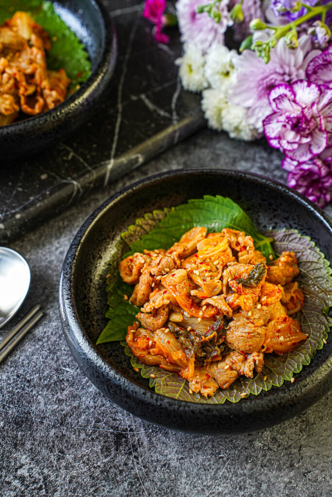 Close up of Pork and Kimchi Stir-Fry over perilla leaves in a black bowl with purple flowers in the background.
