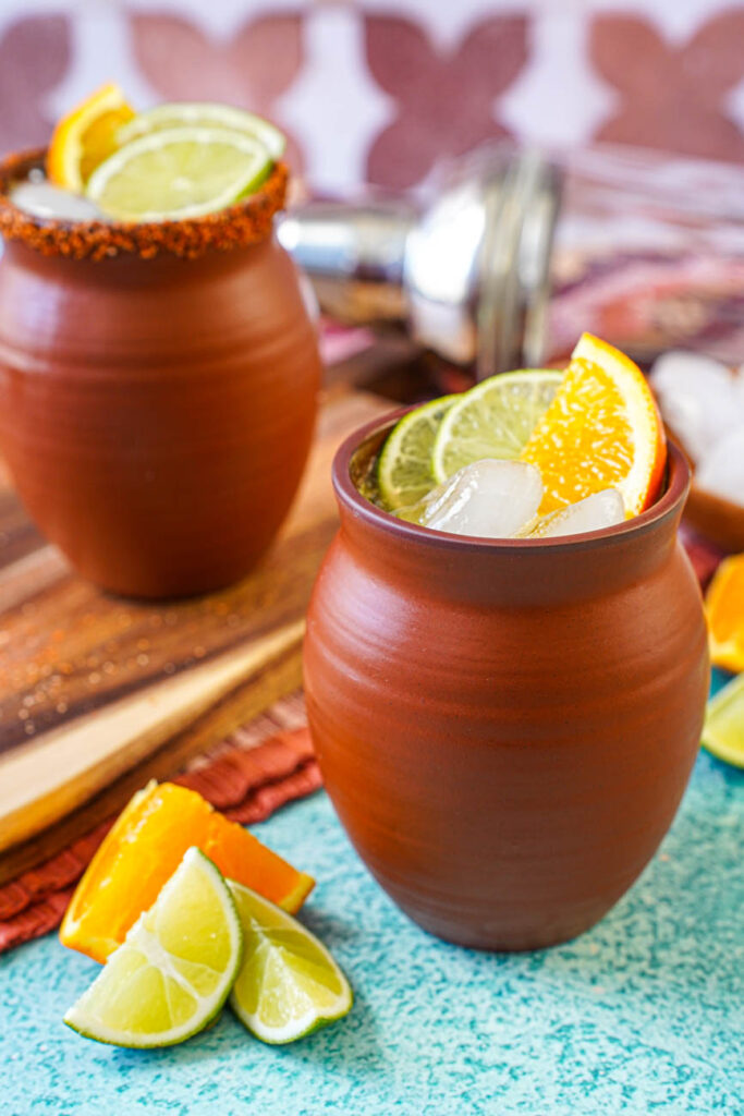 Close up of Cantarito in a clay cup with ice cubes, lime slices, and an orange slice.