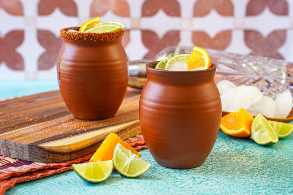 Cantarito in two clay cups next to orange and lime wedges.