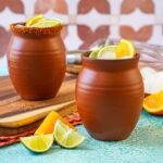 Cantarito in two clay cups next to orange and lime slices.