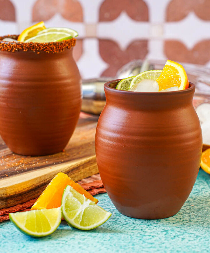 Cantarito in two clay cups next to orange and lime slices.