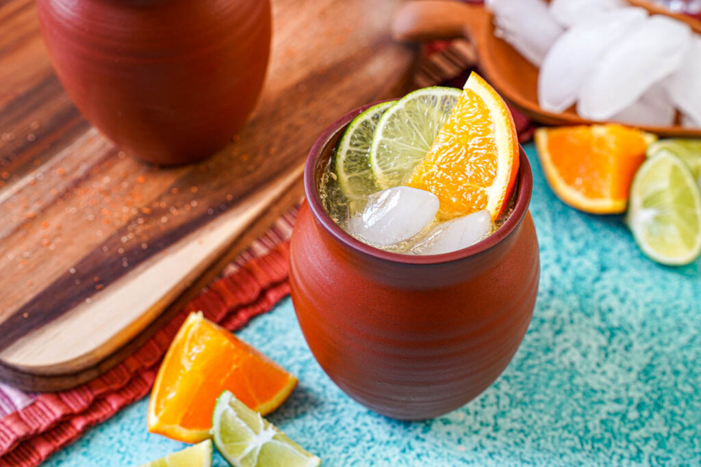 Cantarito in a clay cup with ice cubes, lime slices, and an orange slice.