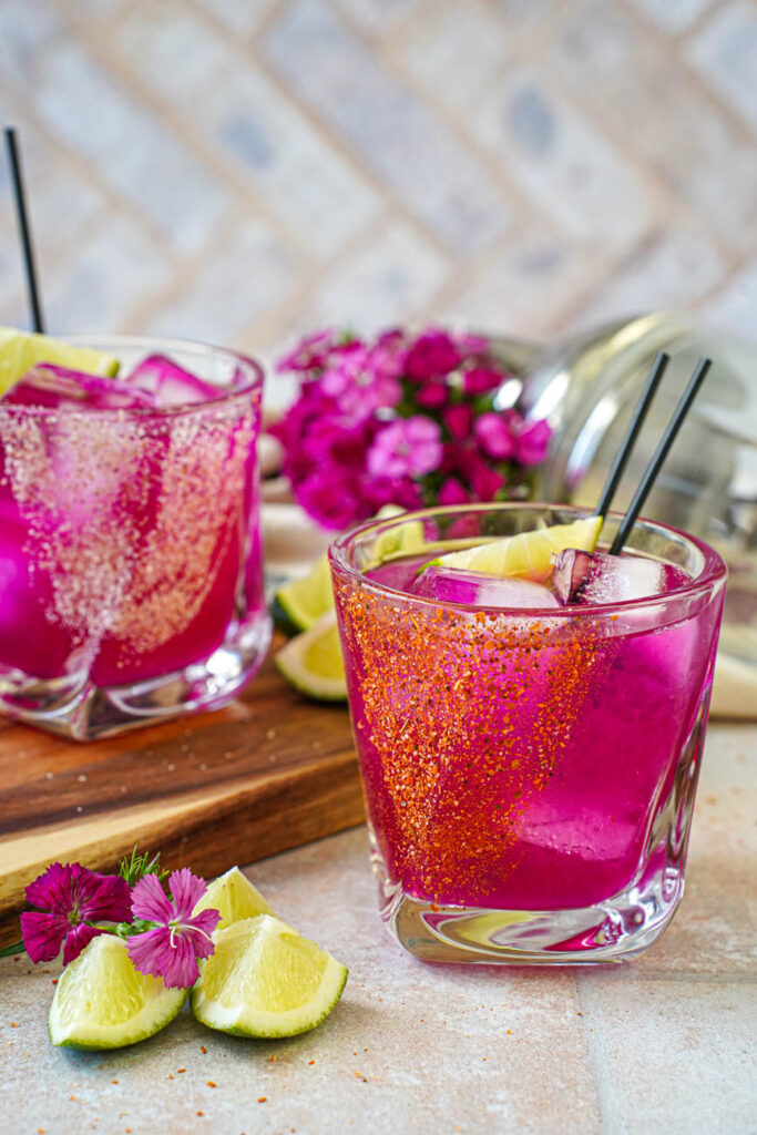 Side view of Prickly Pear Margarita in two glasses with purple flowers and lime wedges in the background.